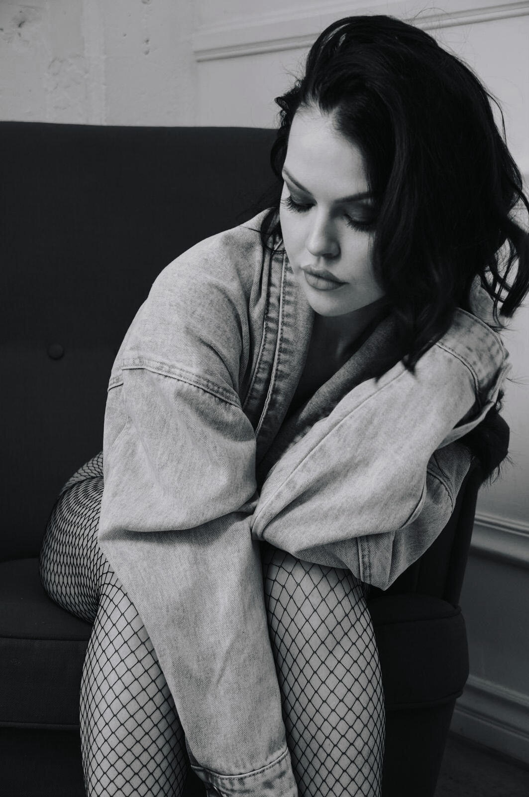 Heather Grace Hancock Black and white portrait of a woman in a denim jacket and fishnets, looking down thoughtfully.